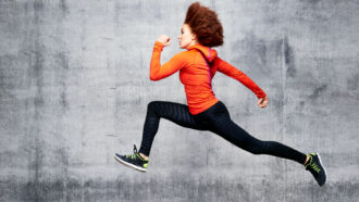 a young Black woman wearing athletic wear, including an orange jacket, black pants and sneakers, sprints across a gray backdrop