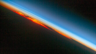 Earth's silhouette against the setting sun is outlined with the reddish brown haze of the lower atmosphere and bluish glow of the upper atmosphere
