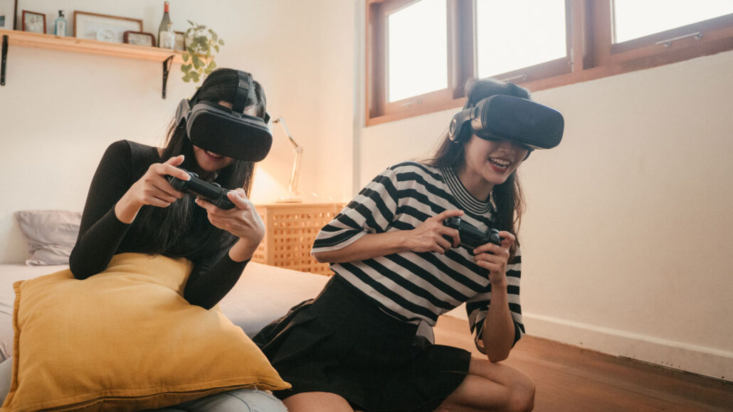 two girls wearing VR headsets and holding handheld controllers sit on the floor and smile as they play a video game