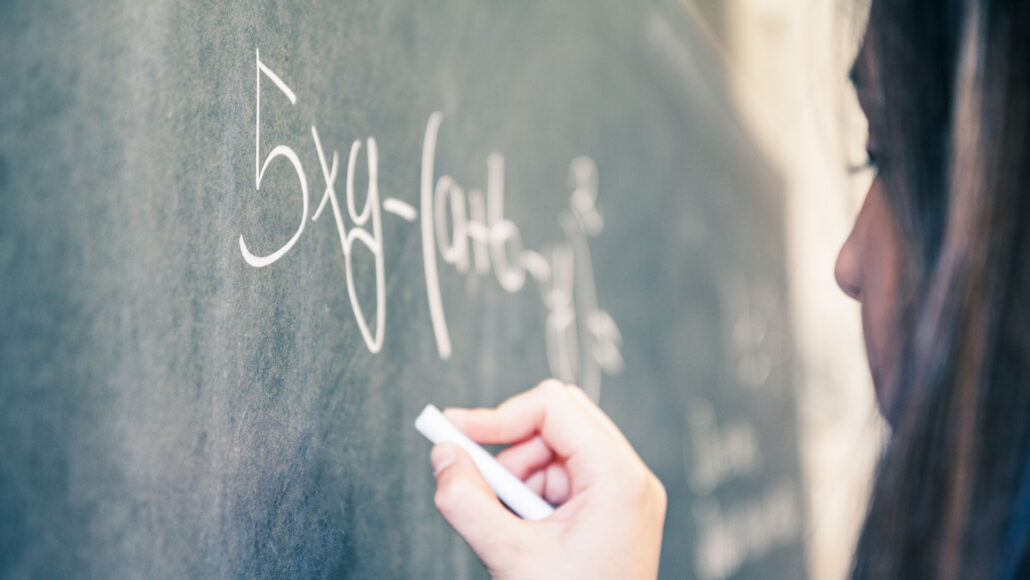 a teen girl holds a piece of chalk up to a chalkboard that has an equation containing "5xy" written on it