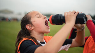 a girl wearing a jersey out on a soccer field squirts water into her mouth from a black plastic bottle