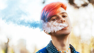 a photo of a teen with a short blue and pink dyed hair exhaling a plume of smoke