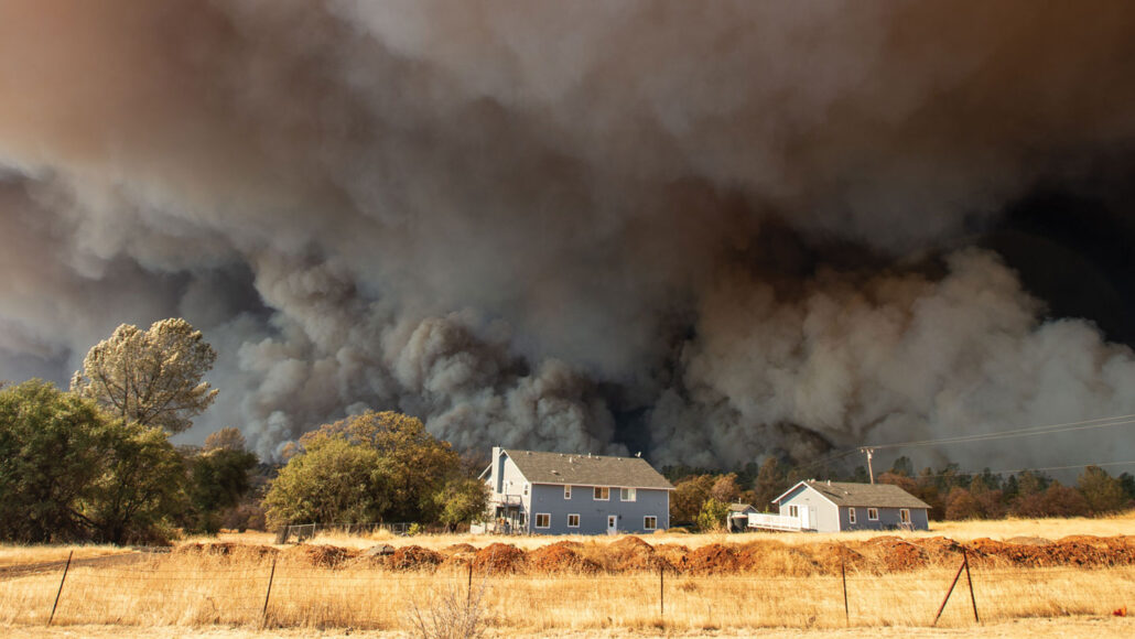 A photo of a yellow pasture with and farm in the distance. Behind the buildings heavy dark smoke clouds fill the sky