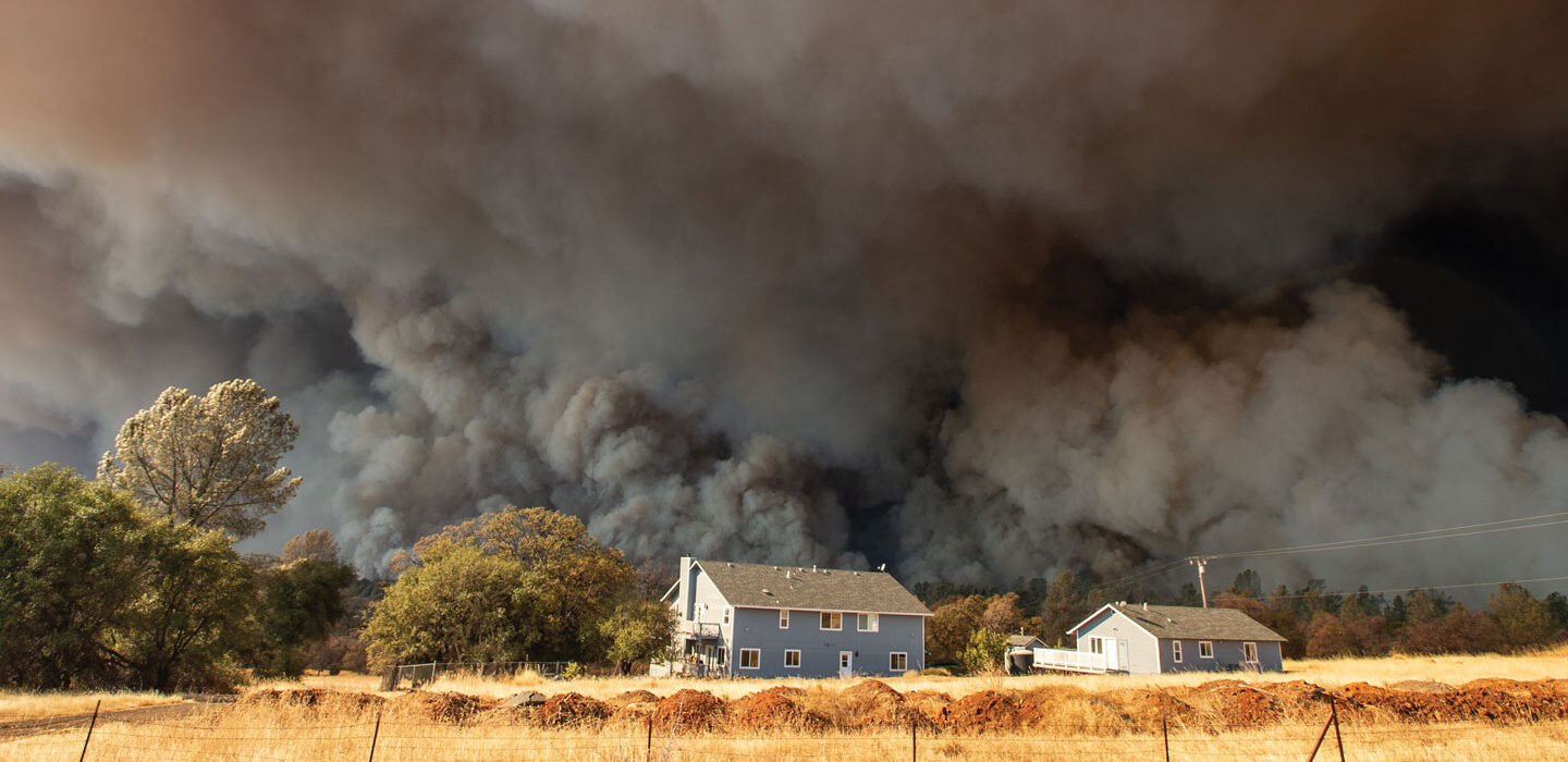 A photo of a yellow pasture with and farm in the distance. Behind the buildings heavy dark smoke clouds fill the sky