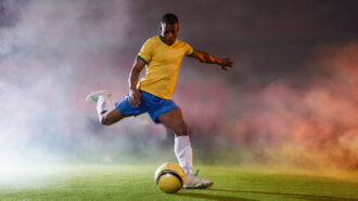 a man wearing a soccer uniform and cleats is about to kick a yellow soccer ball across a pitch