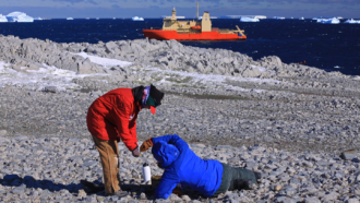 a photo of two researchers collecting samples from a rocky shoreline and a large research ship in the distance at the horizon