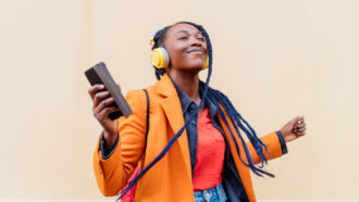 a woman wearing an orange coat, jeans and yellow headphones smiles as she listens to music