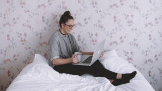 A photo of a young woman drinking from a mug, sitting on her bed, and using a laptop. Floral wallpaper is on the wall directly behind her.