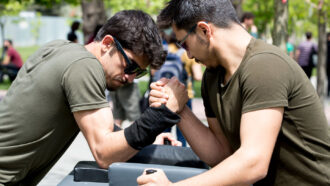two young men wearing sunglasses arm wrestle at a table outside