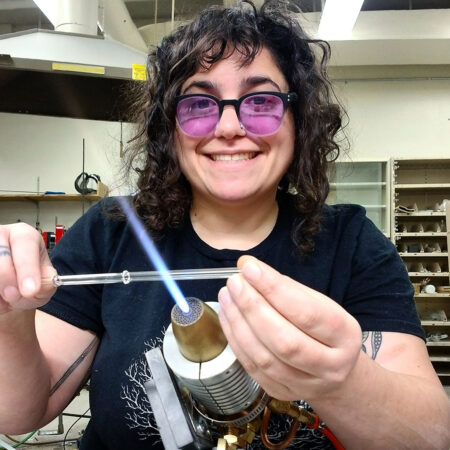a photo of Corinne Guerra, a smiling woman with dark curly hair and purple lensed glasses, heating a small glass pipe 