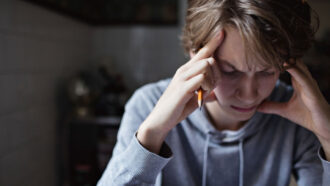 a blonde boy in a grey sweatshirt holds his head in his hands and frowns down at something on his desk