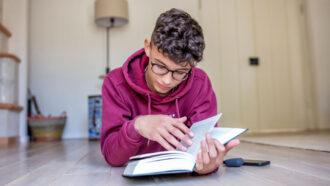 a teen wearing glasses and a red sweatshirt lies on a hardwood floor to read a book in front of them
