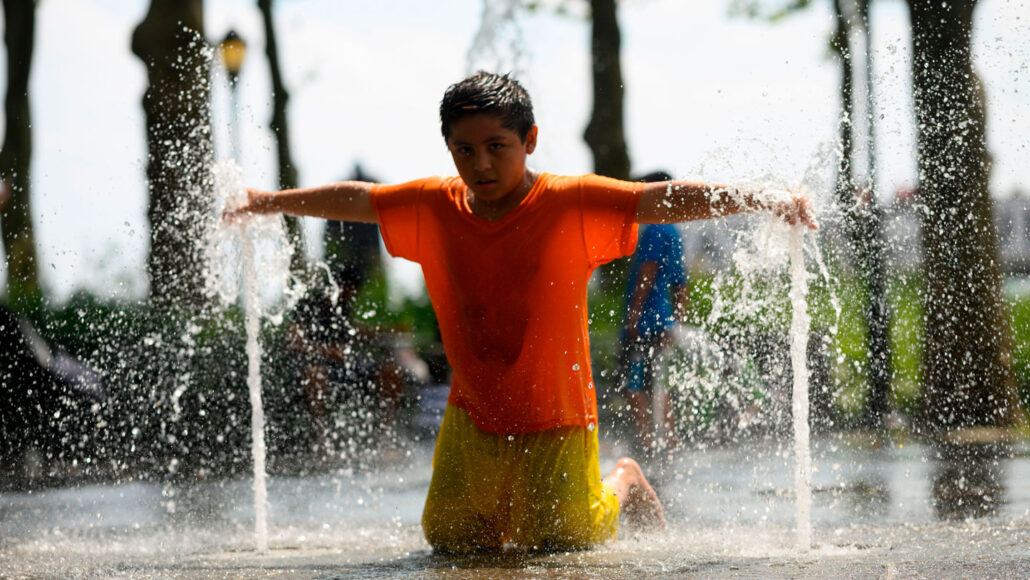 a boy wearing a soaked t-shirt and pants kneels in a fountain and holds his hands over the spouting water