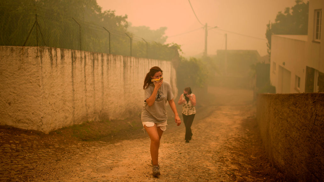 a photo of two girls walking on a dirt paved road while covering their mouths and noses with cloth. The air is hazy and the sky is orange.