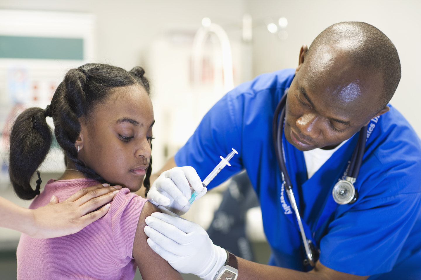 A man wearing blue scrubs injects a vaccine into a teen girl's arm