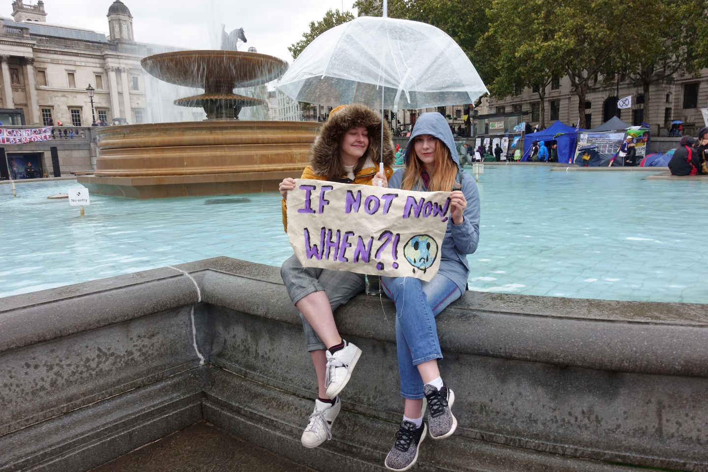 two teen girls sit on the edge of a fountain holding an umbrella and a sign that reads 'if not now, when?!'