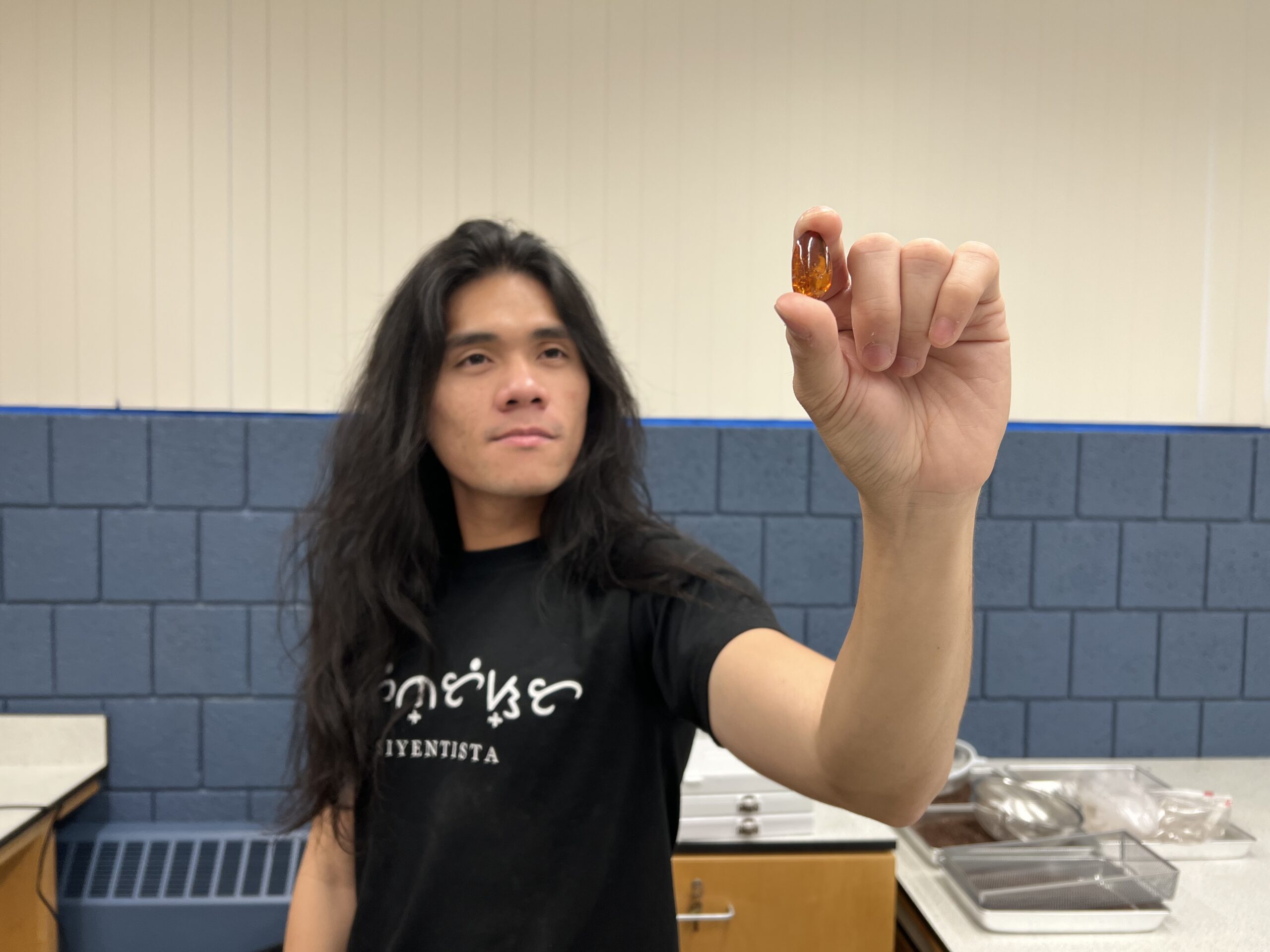 a young man with long black hair wearing a black shirt stands in a lab and holds a tardigrade fossil between this fingers