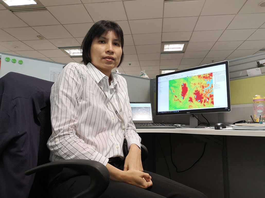 a photo of a woman sitting in front of a computer in a cubicle. The computer monitor shows a map. Teo is wearing a white dress shirt with stripes, she has bangs and shoulder length black hair and is looking at the viewer.