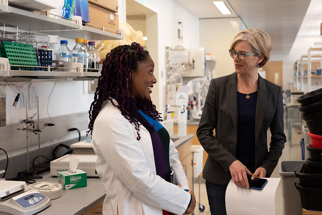 A photo in a lab setting of two women talking. On the left the black woman has thin braids just below her shoulders. She is wearing a black blue and purple dress with a lab coat. An older white woman is to the left. She has glasses and short blond hair. The two women are smiling at each other.
