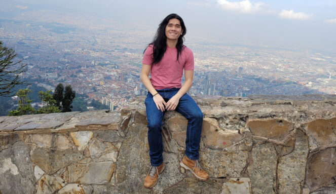 a young man with long black hair and wearing a red shirt and jeans sits atop a stone wall with a cityscape below