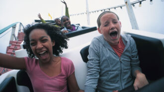 two girls ride in a roller coaster cart flying downhill, one with her hand in the air and the other closing her eyes as she screams
