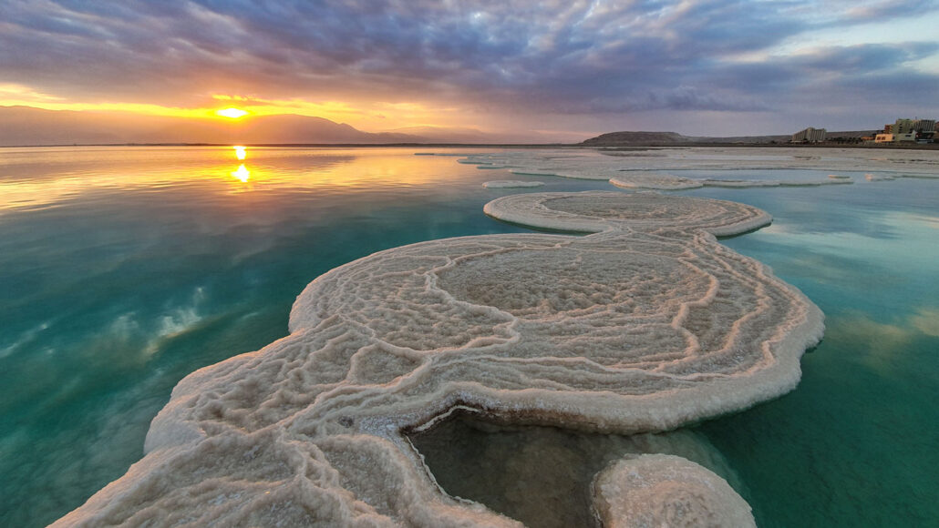 circular patches of salt crystal poke out above the blue green water of the Dead Sea at sunset
