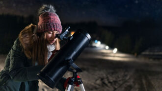 a teen girl in a hat, scarf and coat looks through a backyard telescope on a starry night