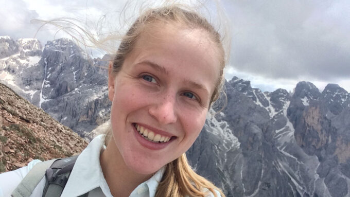 a smiling blonde woman takes a selfie at the top of a mountain under an overcast sky