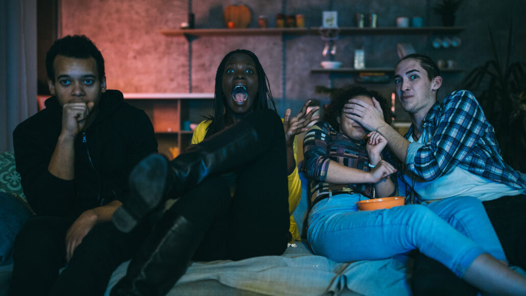 four teenagers sit on a couch in a darkened room, screaming or covering their eyes in response to something unseen on a TV
