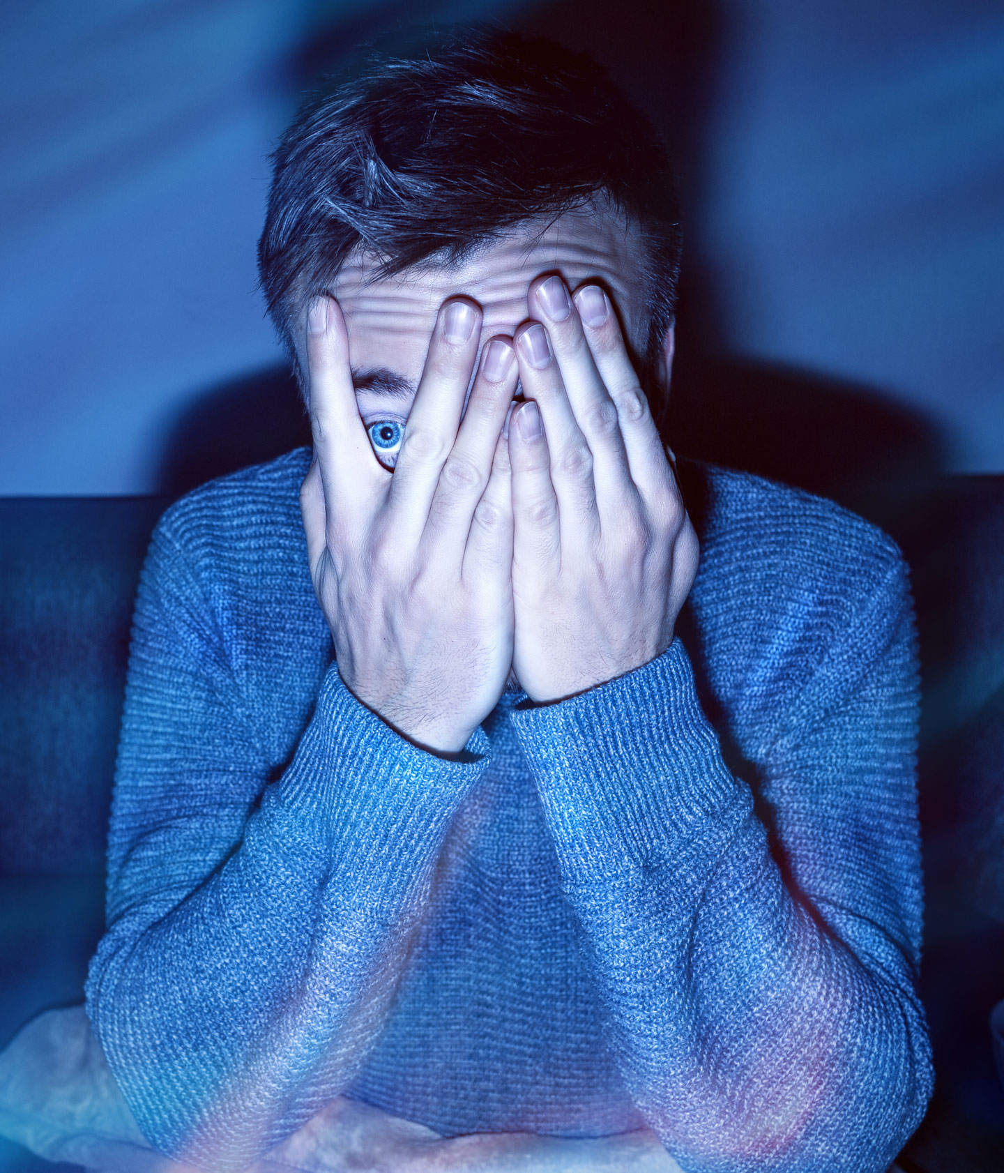 a teen boy whose face is illuminated by a TV screen peeks through his fingers with one wide eye