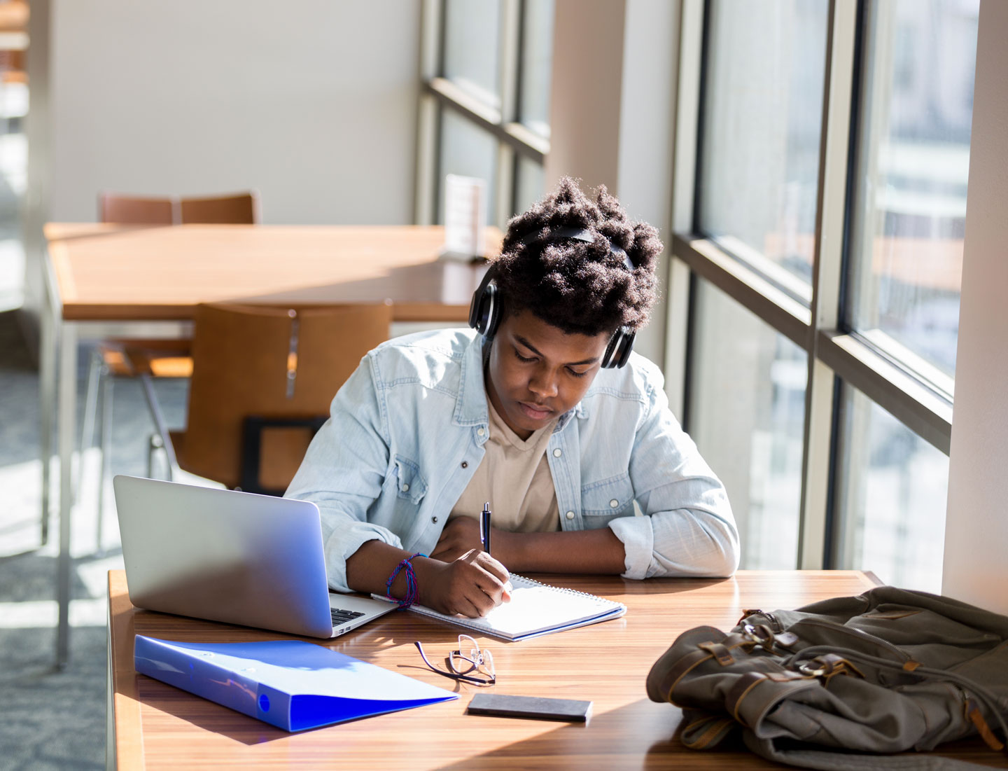 a teen boy sits at a desk beside a window with a notebook, laptop and binder