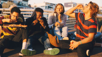 four teen boys sit on a sunny roof and eat pizza