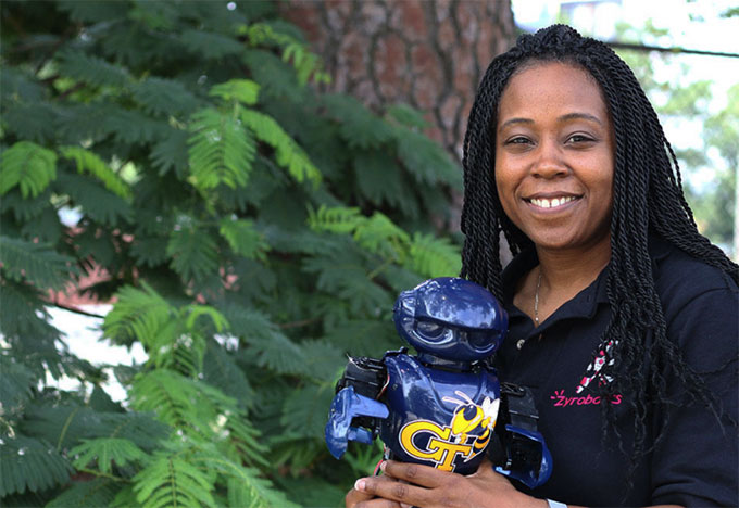 Ayanna Howard smiles at the camera. She has long hair in twists and is holding a small blue robot with the Georgia Tech logo on it.