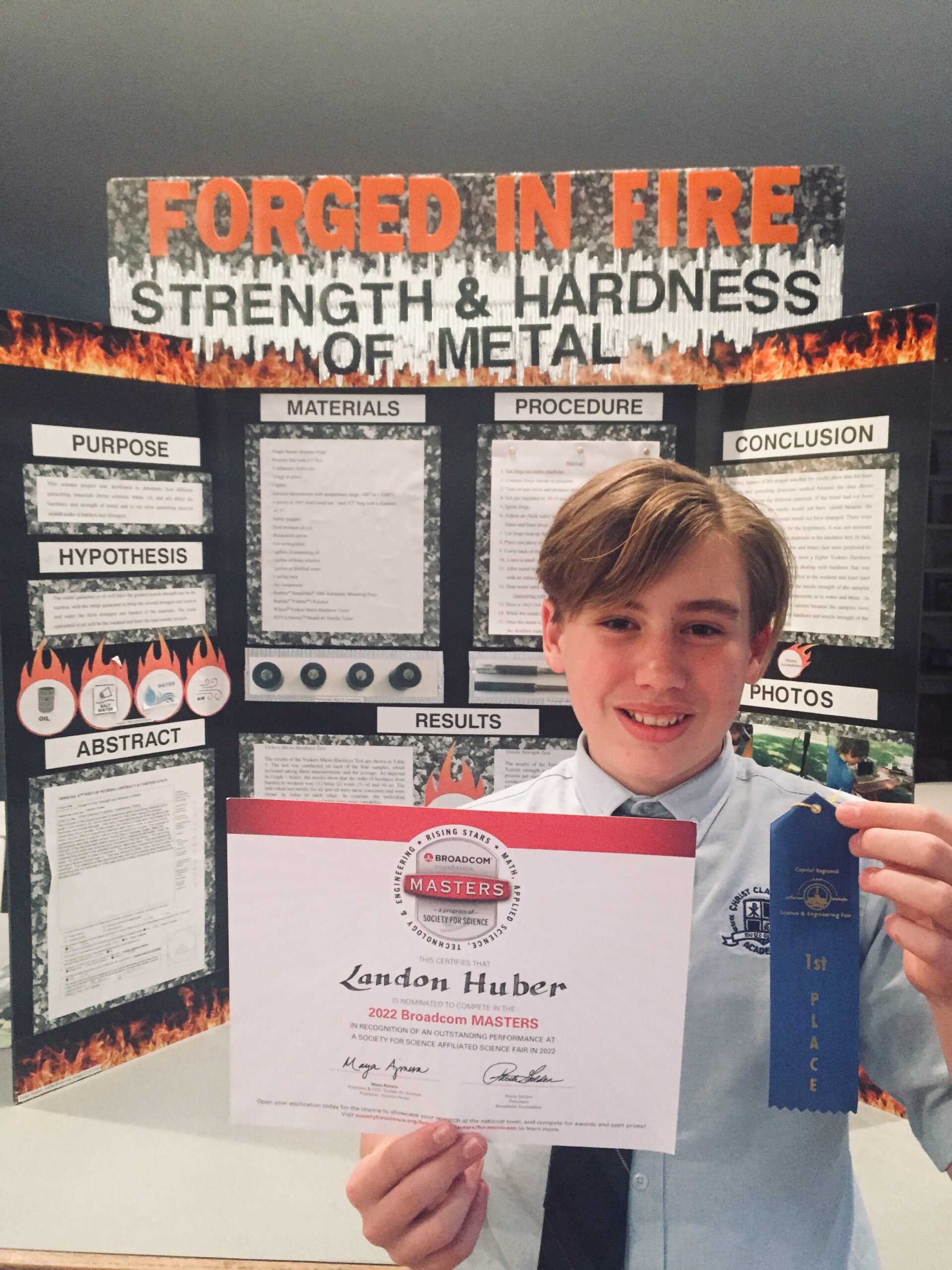 a blonde boy stands in front of a project poster titled 'Forged in Fire' and holds up a blue ribbon and certificate