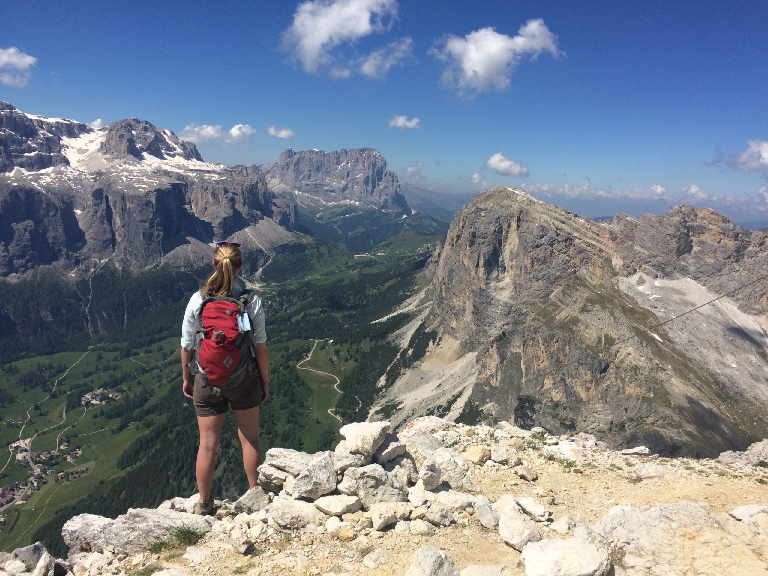 a woman wearing a red backpack faces away from the camera to look out from a mountaintop at a mountain range under a blue sky