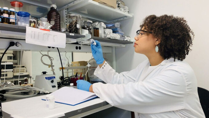 a woman wearing a white lab coat holds up a vial to inspect while sitting at a lab bench