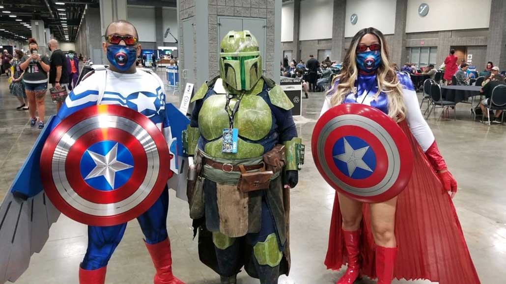 Tina Saey stands between two versions of Captain America. She is wearing green Mandalorian armor and a green Mandalorian helmet.