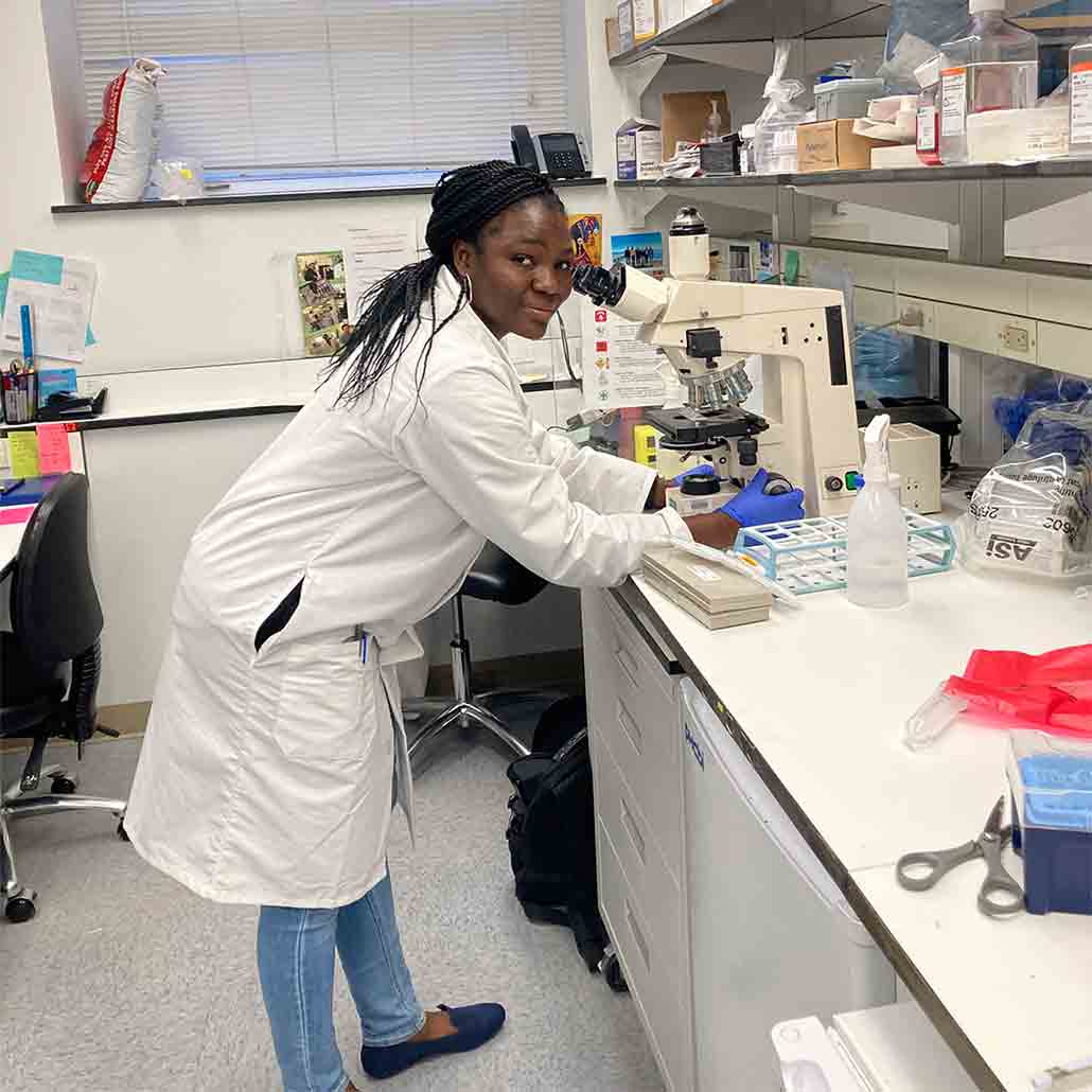 a photo of Oyebola Oyesola, a smiling lady with dark skin and braids, bending over a microscope in her lab