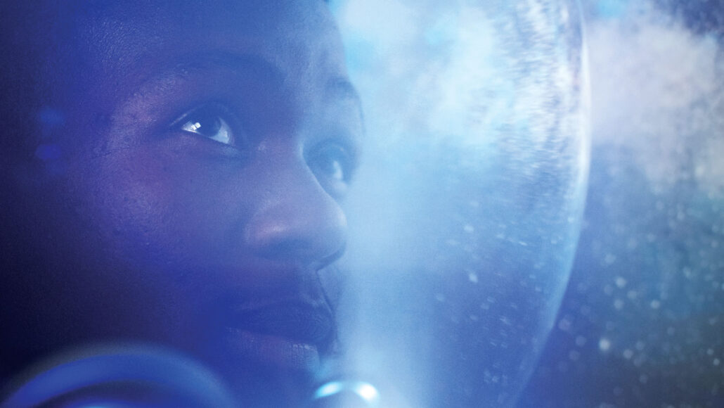 A Black man looks out of an astronaut helmet with outer space in the background