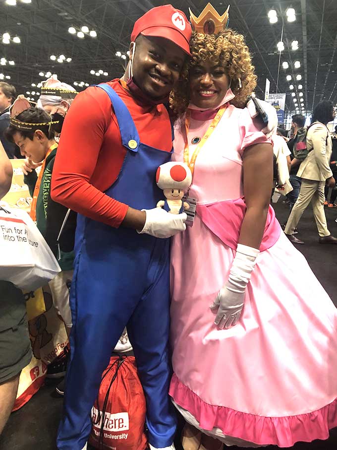 Two smiling Black cosplayers, one dressed as Mario the other as Princess Peach. Mario is holding a small Toad stuffy.