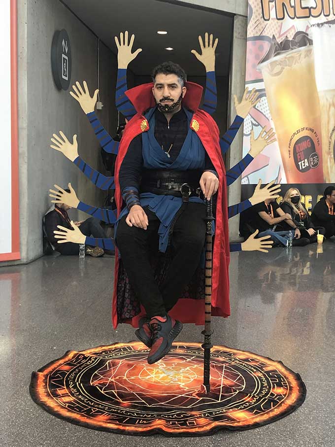 A man appears to float in midair above a carpet patterned with an arcane circle. He has 10 cardboard hands projecting from behind his back. He's wearing the costume of Dr. Strange.