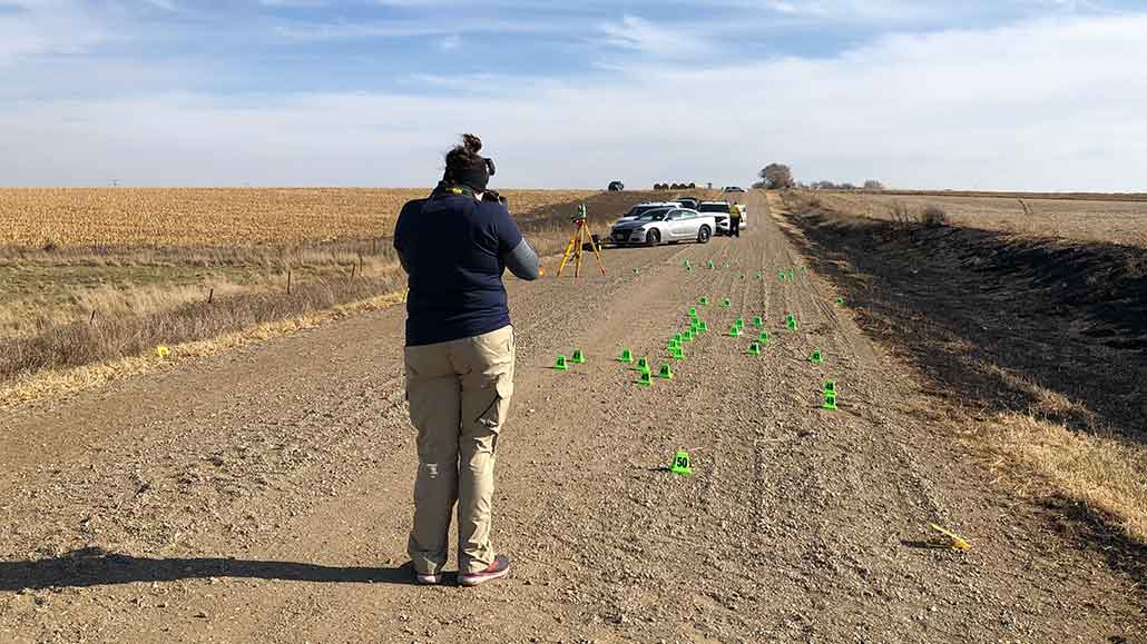 a photo showing Samantha Hayek facing away down a dirt road speckled with green evidence flags. She is taking photos of the scene. 