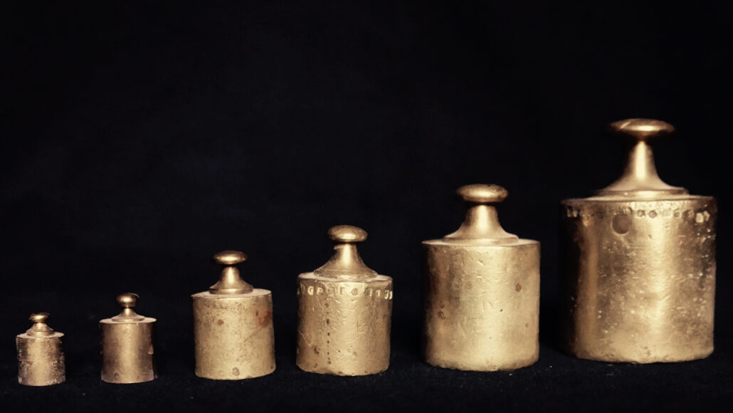 A photo of six old fashioned golden weight in a line from the smallest weight to the left and the biggest weight on the right, all on a black background