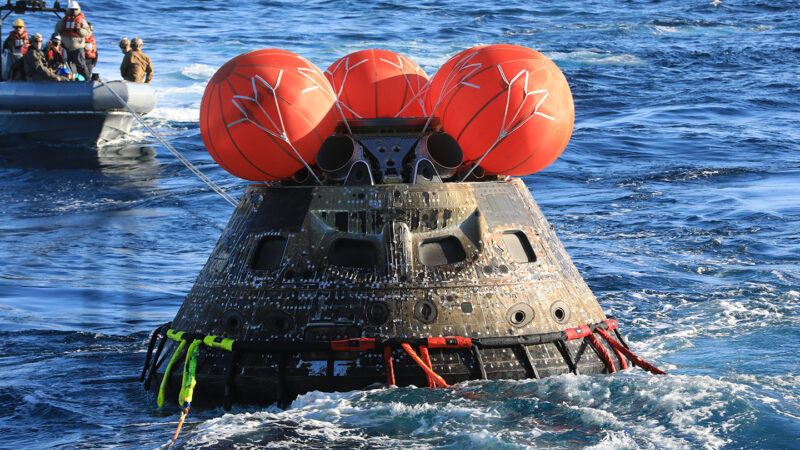 the uncrewed space capsule Orion floating in the Pacific Ocean, with round red airbags atop it as a crew in a boat in the background retrieves the capsule