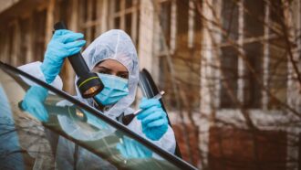 a photo of a woman in a clean suit wearing a face mask and blue gloves. She is inspecting the edge of a car windshield with a light and a brush, looking for fingerprints