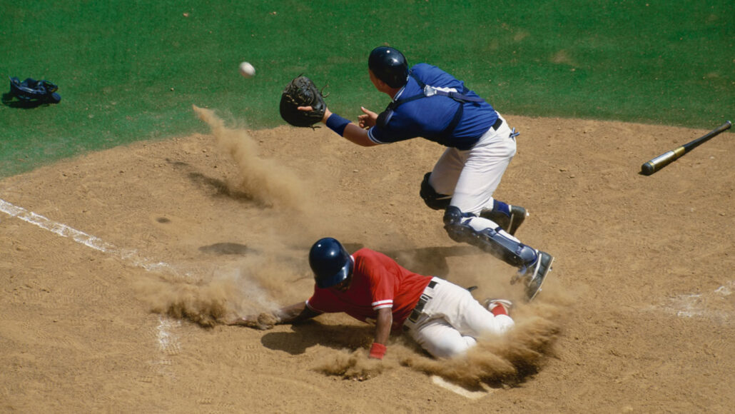 a baseball player slides into home base, kicking up dust, as the catcher reaches for an incoming baseball