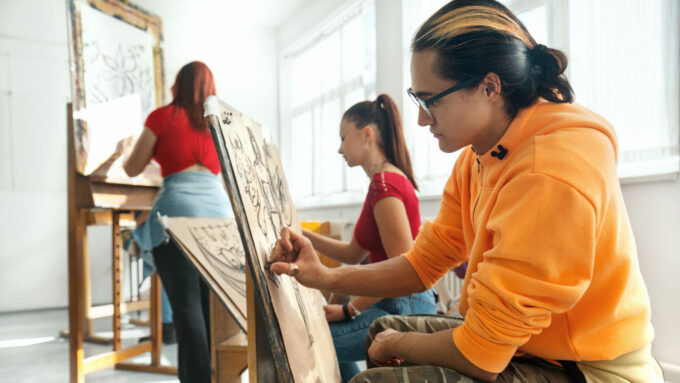 a boy in an orange sweatshirt and cargo pants draws on an easel with charcoal
