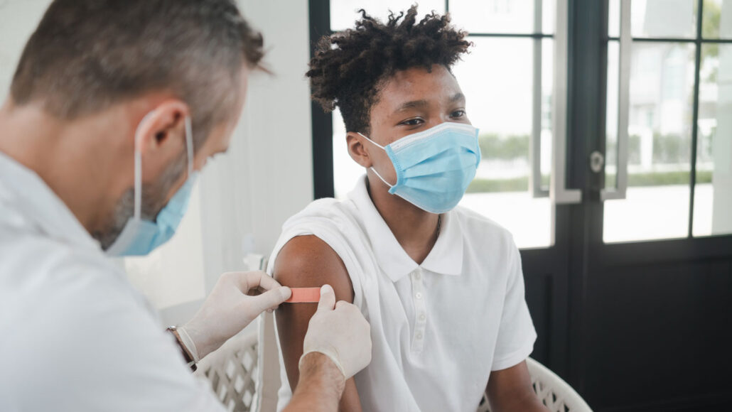 a teen boy sits on an exam table while a nurse pits a band-aid on his upper arm