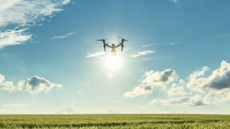 a drone flies over a field under a blue sky, its silhouette backlit by the sun