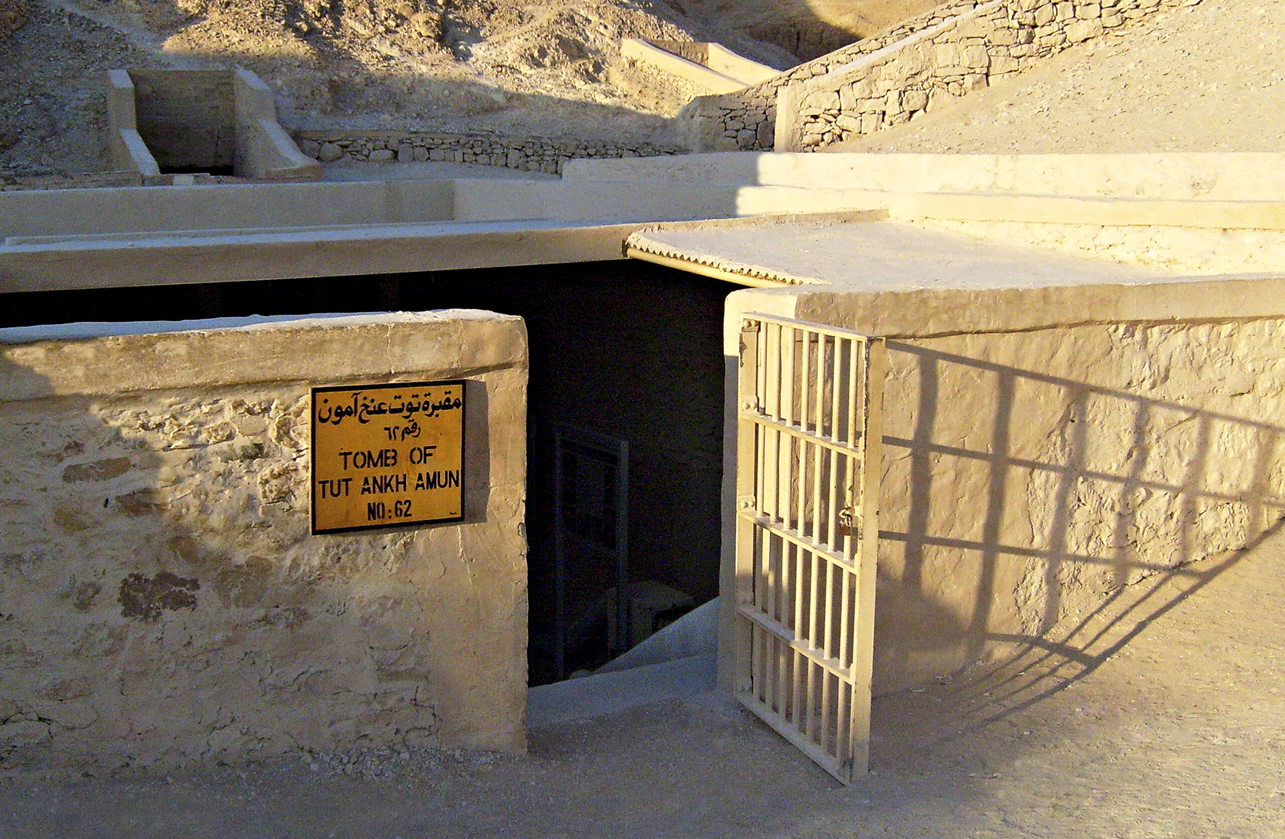 a wall and gate surround a set of stairs leading down into the ground of a desert; a plaque on the wall reads 'tomb of tutankhamun'